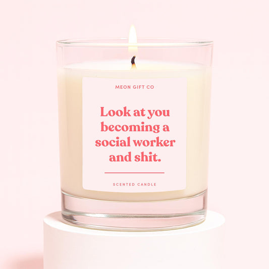 Clear-glass scented candle with flame; label reads 'Look at you becoming social worker and shit.'